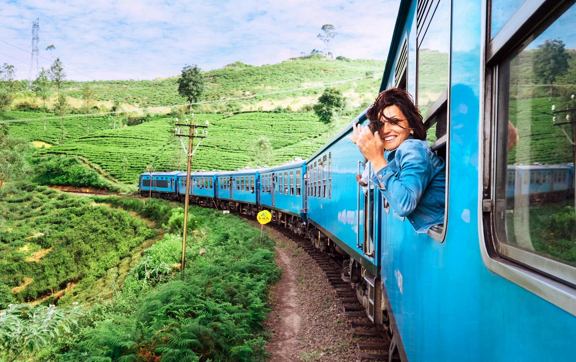 A smiling woman looks out from a train window as it travels through a hillside location covered in greenery